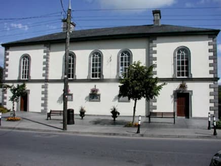 Ein zweigeschossiges weiß verputztes Gebäude mit Ecksteinen aus Werkstein und hohen Rundbogenfenstern, Bänken und Blumenkörben an der Straßenfront.