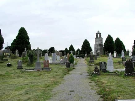 Ein Kiesweg durch einen Friedhof mit aufrechten Grabsteinen und dunklen Nadelbäumen, mit einem hellen Mausoleum unter einem Kreuz.