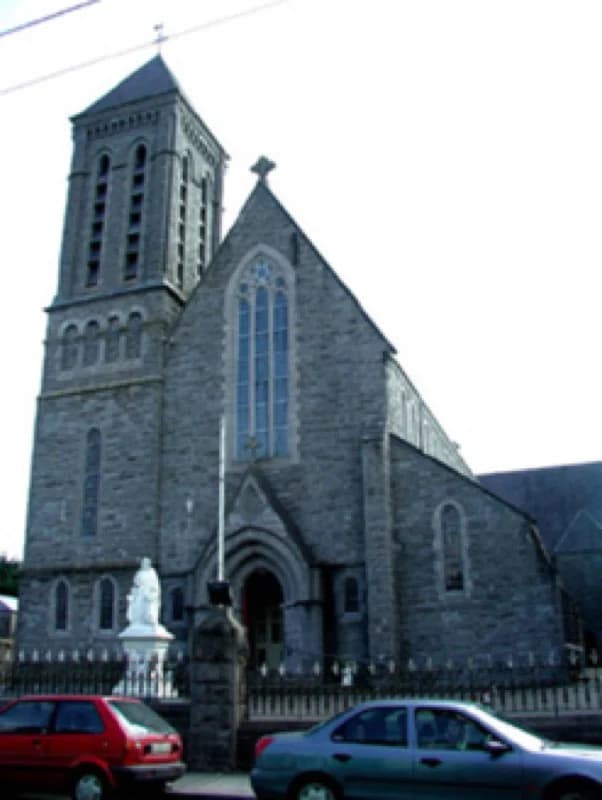 A cut-stone church with a square tower and spire, lancet windows, and a white statue on a pedestal inside the railings.