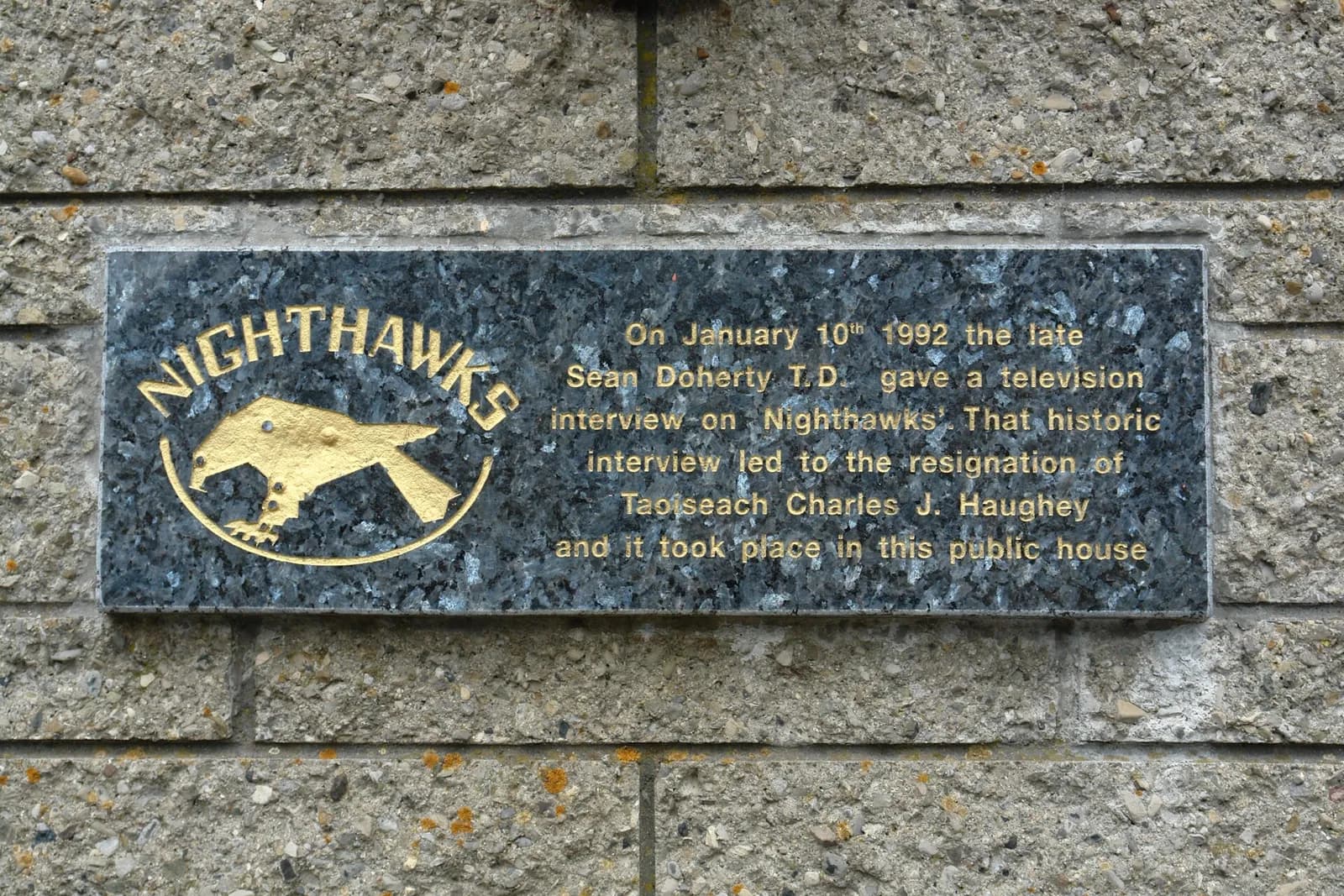 Polierte dunkle Granittafel mit Goldschrift in einer Betonsteinmauer, überschrieben NIGHTHAWKS über einem goldenen Falken, zum Fernsehinterview vom Januar 1992.