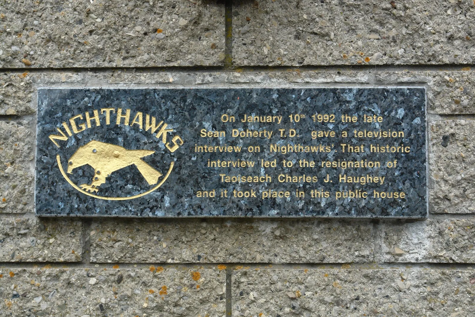 Polierte dunkle Granittafel mit Goldschrift in einer Betonsteinmauer, überschrieben NIGHTHAWKS über einem goldenen Falken, zum Fernsehinterview vom Januar 1992.