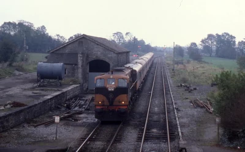 Orange-schwarze Lokomotive mit Wagenzug neben einem steinernen Güterschuppen in Castlerea bei flachem Herbstlicht, Schwellen entlang des Gleises gestapelt.