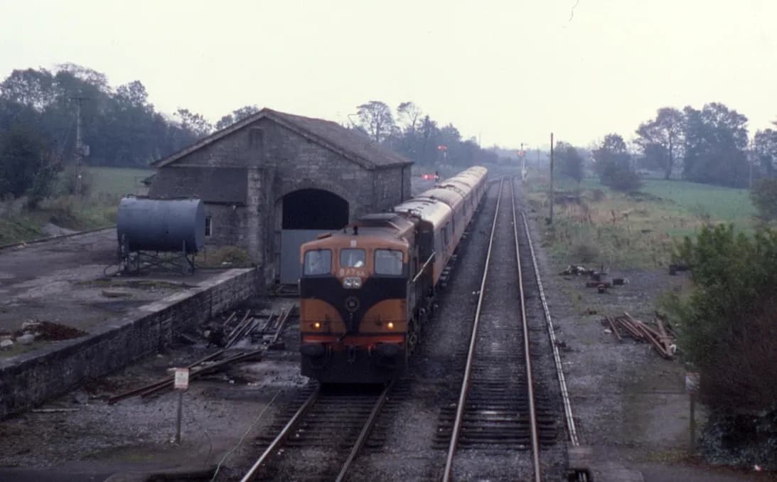 Orange-schwarze Lokomotive mit Wagenzug neben einem steinernen Güterschuppen in Castlerea bei flachem Herbstlicht, Schwellen entlang des Gleises gestapelt.