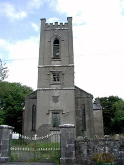 Una chiesa intonacata con torre merlata a tre ordini, finestre a tutto sesto e cancellate in ferro su pilastri in pietra lungo la strada.
